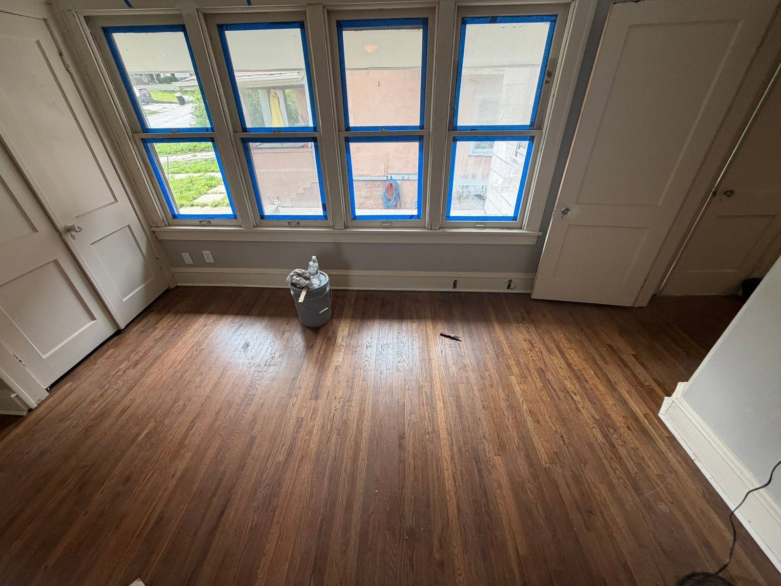 Hardwood floor mid-refinish with windows taped off and a finish can on the floor
