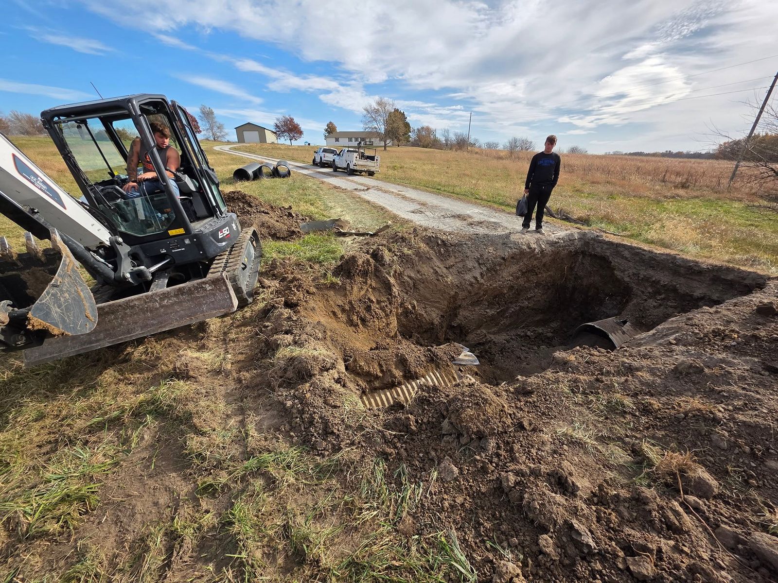 Compact track loader digging out a hole for a culvert, with corrugated pipe staged nearby