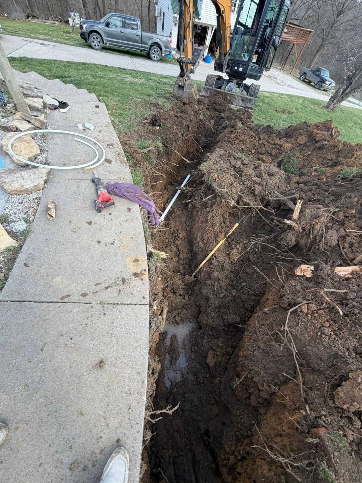 Trench dug along a sidewalk with conduit already laid in and the excavator bucket overhead